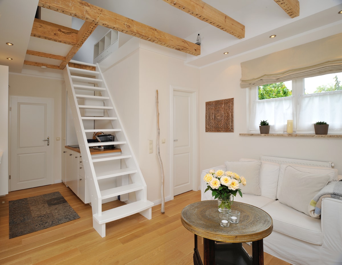 A light-filled living area features a white sofa positioned near a wooden coffee table. A staircase with a minimalist design leads to an upper level. Soft white curtains frame the window, and a decorative wall piece complements the neutral color scheme.