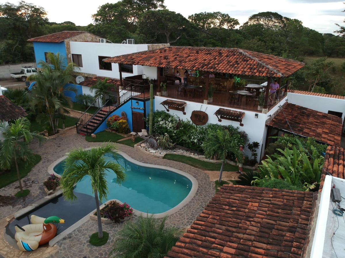 Aerial view of the property showcases a well-maintained pool surrounded by lush greenery and colorful plants. The building features a terracotta roof and an outdoor dining area on a raised deck, providing a panoramic view of the tranquil surroundings.