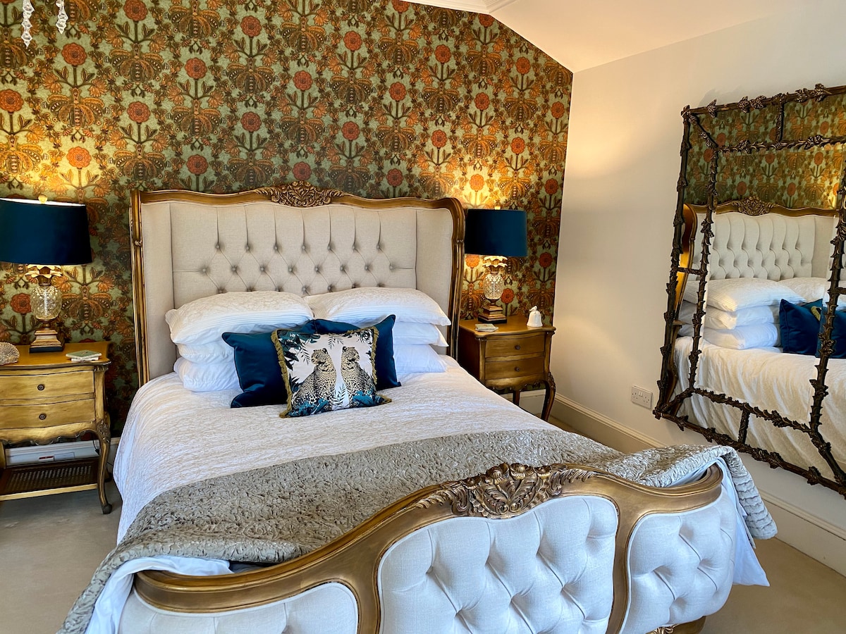 The room features an elegant super king bed with plush linens, set against a detailed floral wallpaper. Two bedside tables with lamps frame the bed, while a vintage mirror reflects the room’s warm tones. A decorative cushion adds a pop of color.