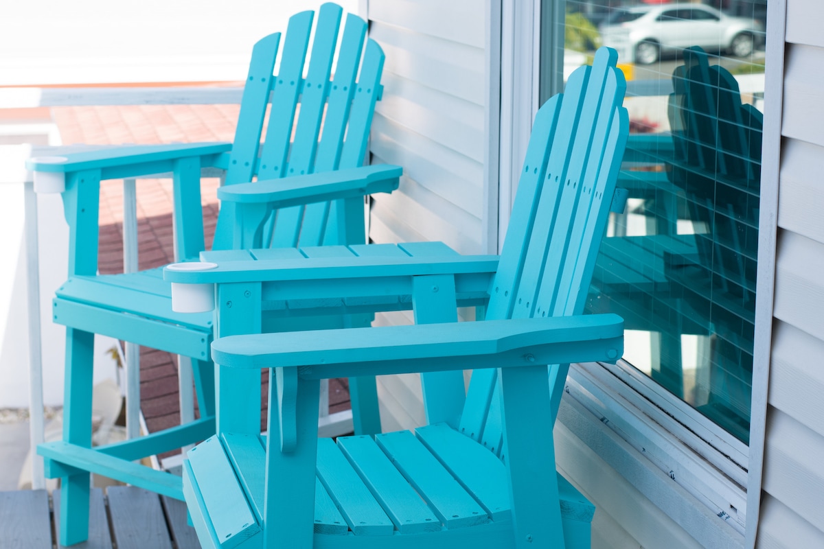 Two turquoise Adirondack chairs are positioned on a balcony, positioned beside a large window. The bright color of the chairs contrasts with the neutral tones of the building's exterior. Reflections of the surrounding area can be seen in the window.