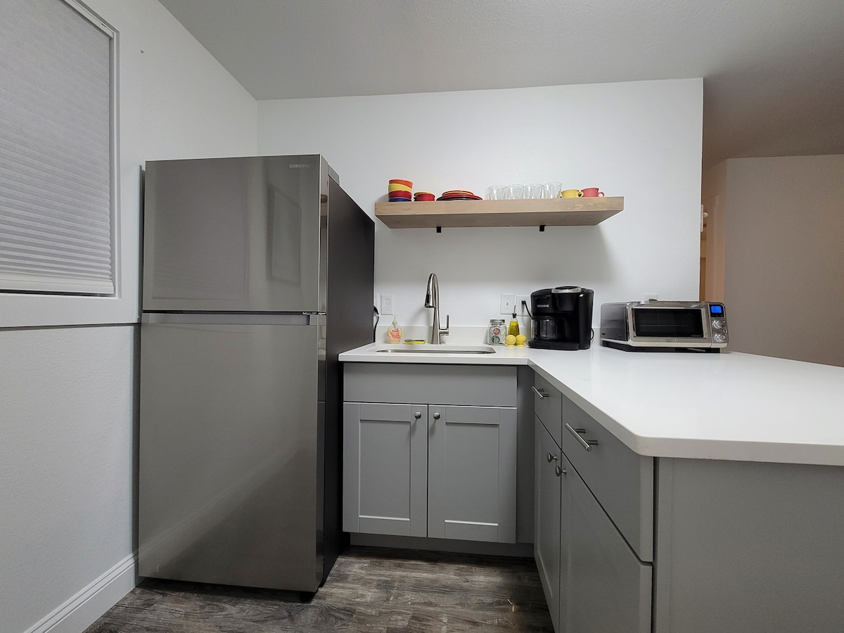 The kitchen area features a modern stainless steel refrigerator and a sleek countertop with a sink. Open shelving displays colorful dishware, while a microwave and coffee maker are positioned for convenience on the counter. Soft lighting enhances the clean and functional design.