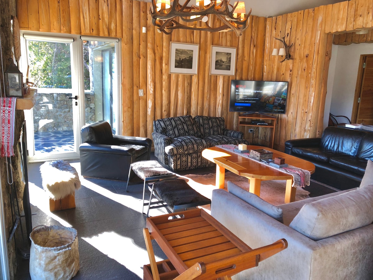 A cozy living area is surrounded by wooden walls and features a mix of seating options, including a black leather sofa and a patterned couch. A wooden table sits in the center, with a TV mounted on the wall, and light streams through glass doors leading outside.