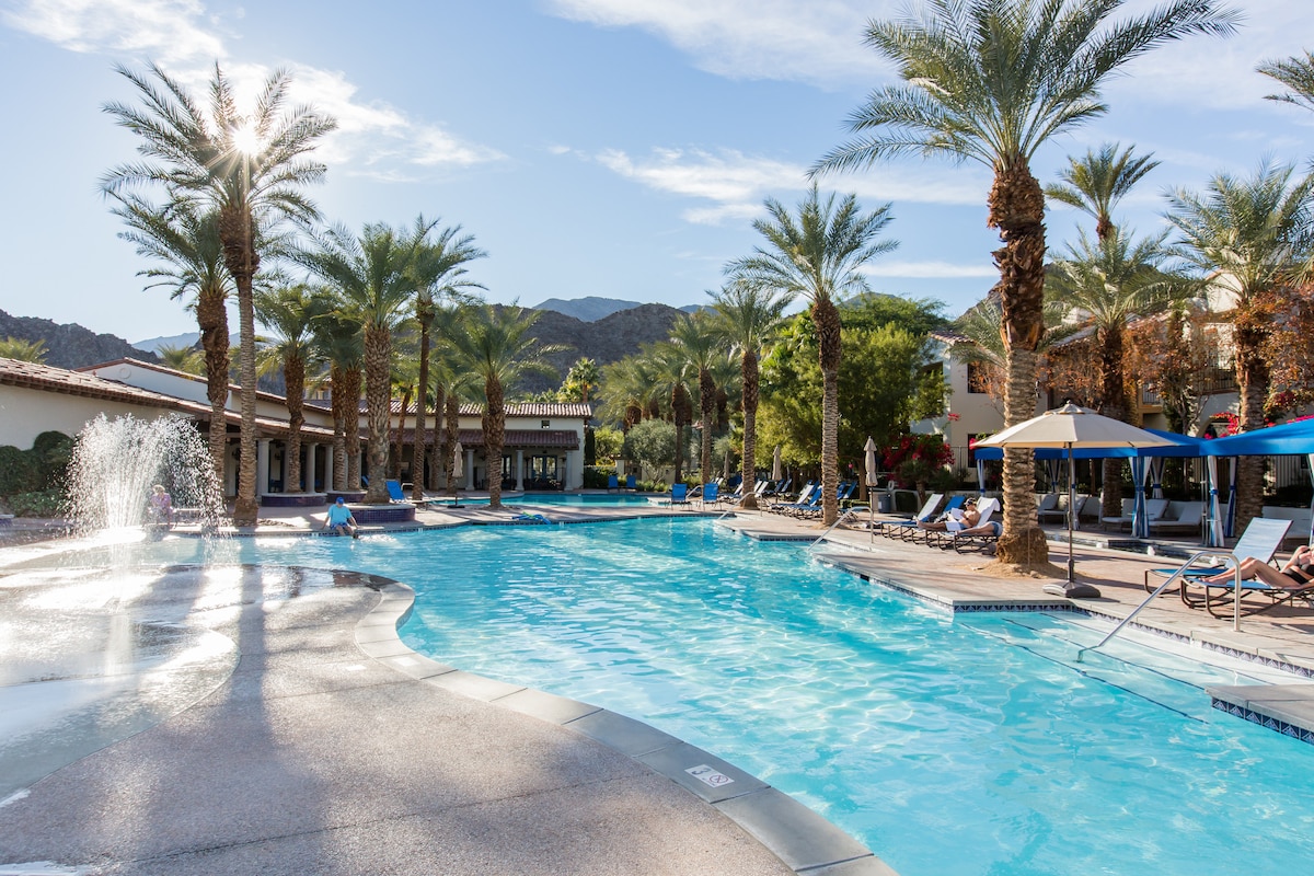 A resort pool area is surrounded by tall palm trees, with lounge chairs positioned along the water. A gentle fountain can be seen, creating a relaxing atmosphere. Scenic mountains are visible in the background under a clear blue sky.