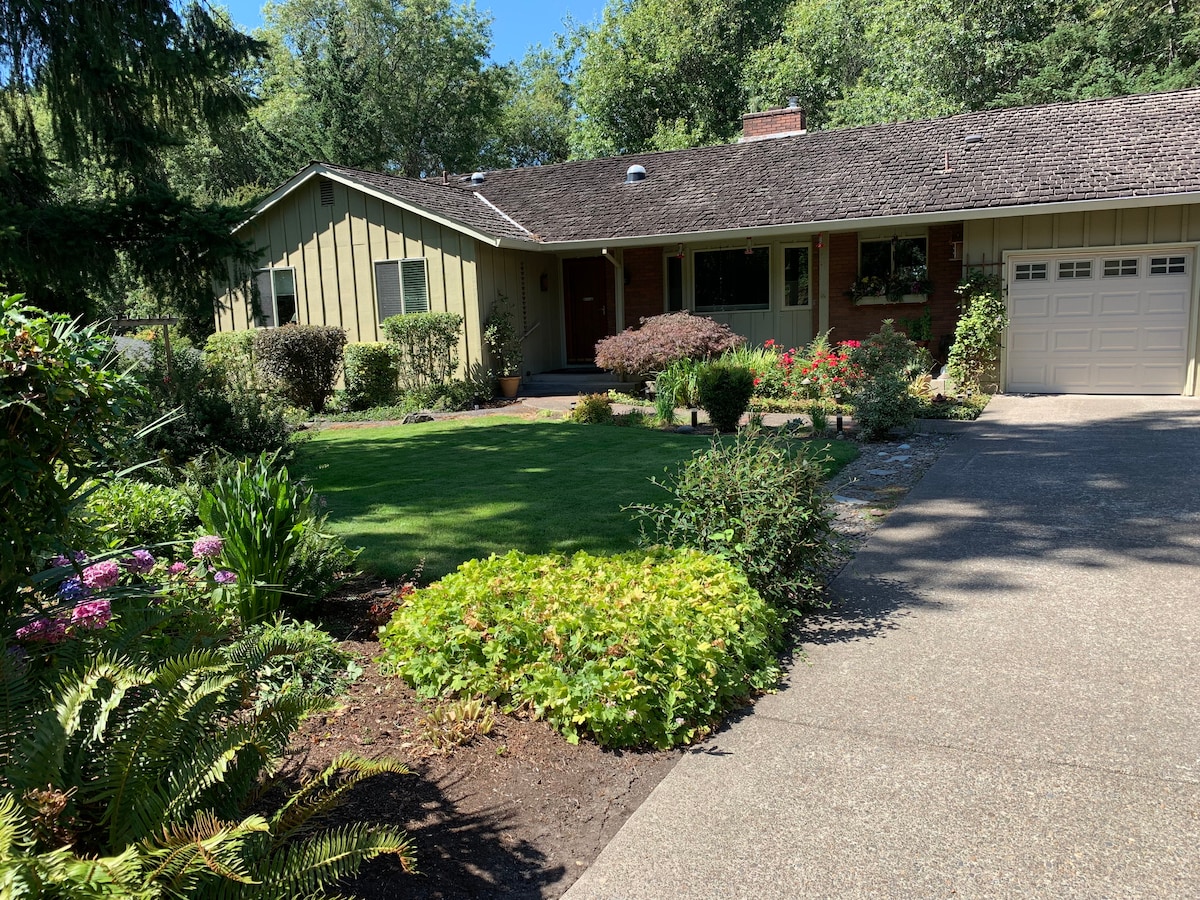 A welcoming exterior view of a single-story home is set amidst lush greenery. A well-maintained garden features various flowering plants and shrubs, while a concrete path leads to the front door. A single garage is visible on the right side of the image.
