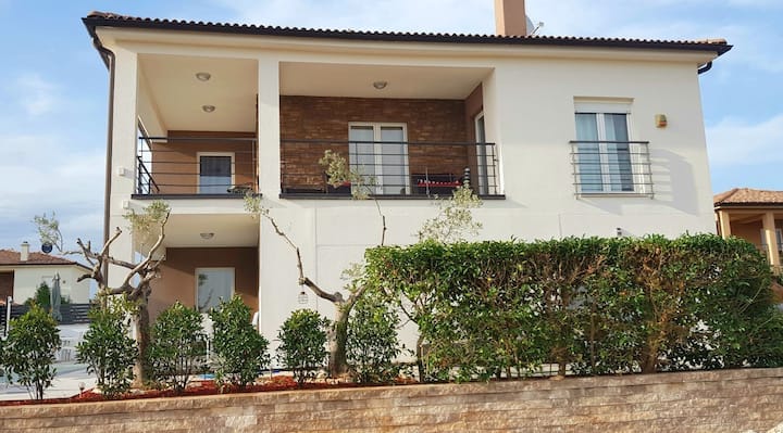 The house exterior showcases a modern two-story design with a combination of light-colored walls and natural stone accents. A spacious balcony is visible on the upper floor, framed by greenery. The surrounding area is landscaped with neatly trimmed bushes, enhancing the overall appeal.