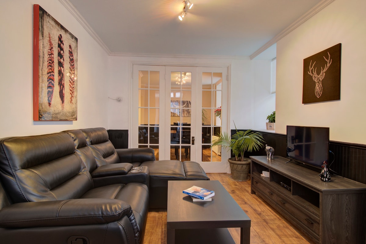 A modern living area features a dark leather sectional sofa, with a sleek coffee table in front. Decorative artwork adorns the walls, and a television is positioned on a dark wood media console. Light enters from glass-paneled doors leading to a separate space.
