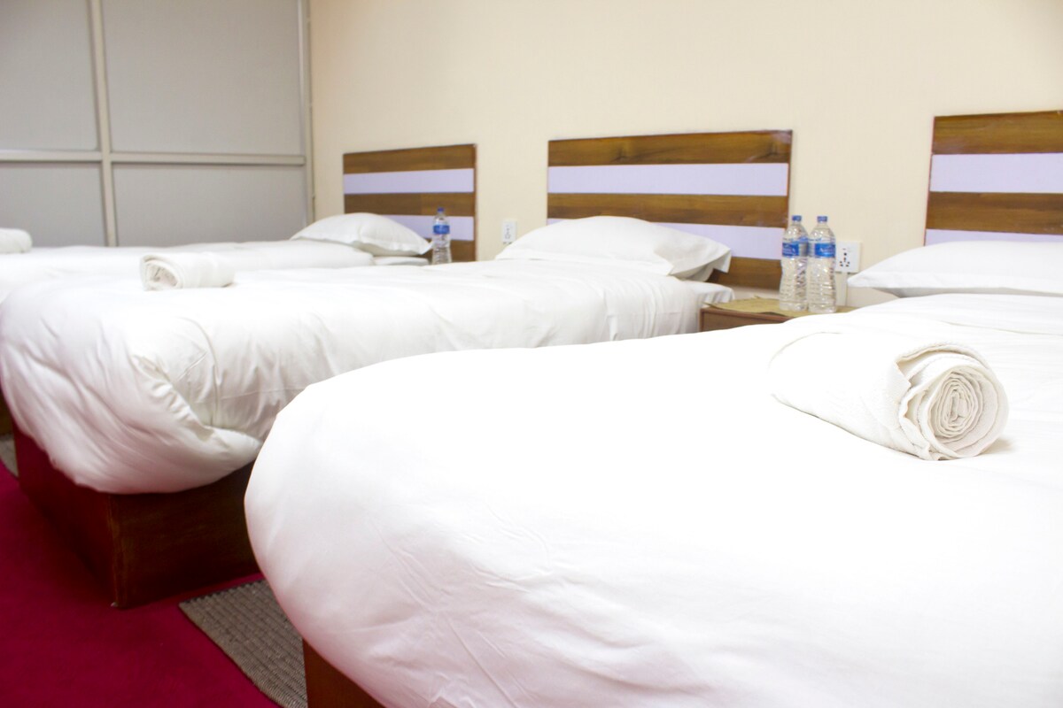 A clean and well-arranged guest room displays three spacious beds, each topped with fresh white linens. Wooden headboards provide a simple touch. Bottled water is positioned on a bedside table, complemented by neatly rolled towels placed on the bed covers.