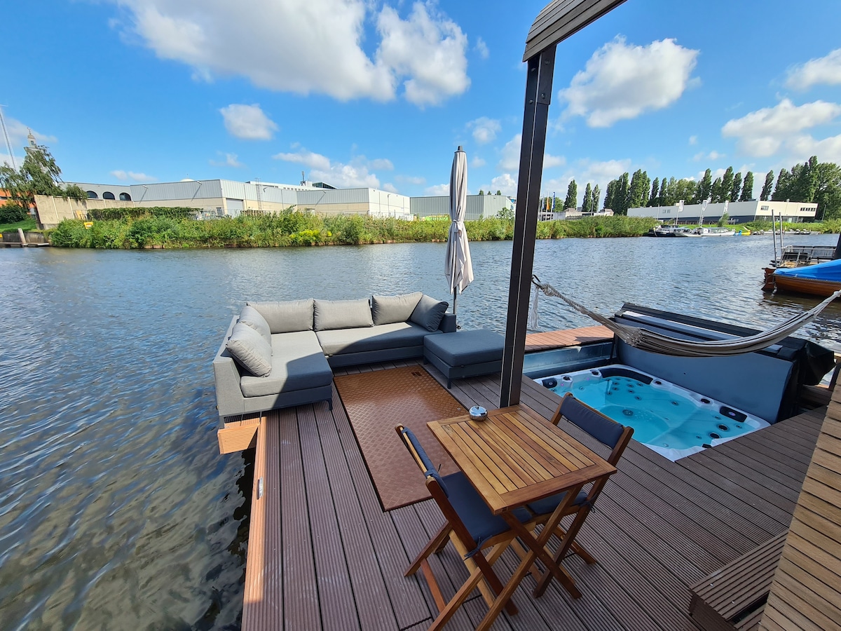 A spacious outdoor terrace features a comfortable gray sectional sofa arranged around a wooden table. A parasol stands beside the seating area, while a jacuzzi is visible nearby, inviting relaxation by the water's edge. The serene river stretches in the background under a clear sky.