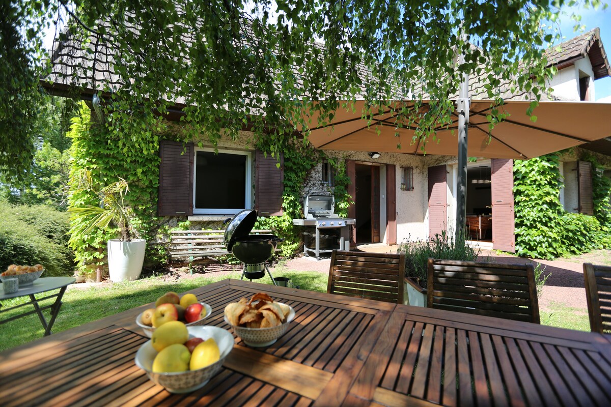 A spacious outdoor dining area features a wooden table set with a bowl of apples and pastries. In the background, a house is partially shaded by a large tree, with a sunshade providing covering over a barbecue area and lush greenery surrounding the property.