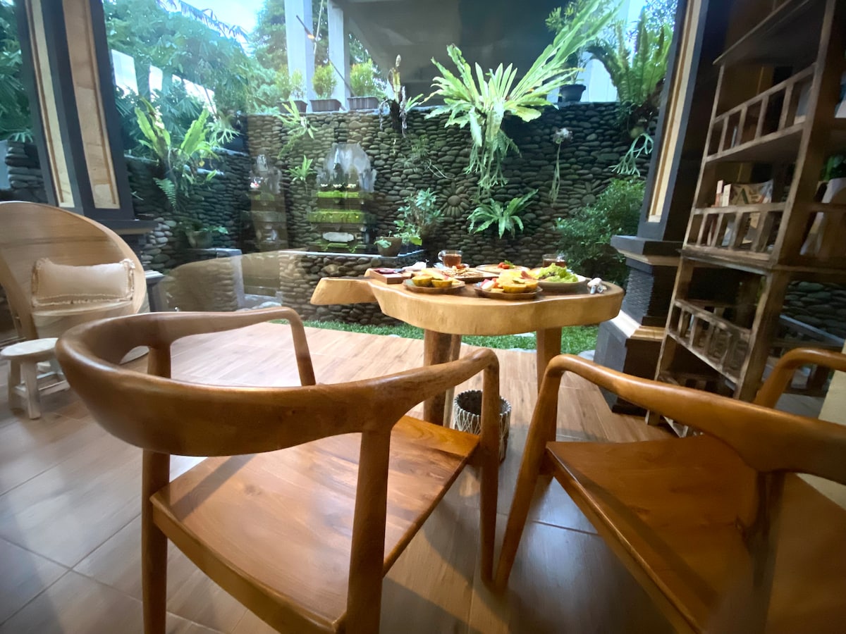 A dining area features natural wood chairs positioned around a round table. A selection of food is displayed on the table, with a lush green garden visible through large windows, highlighting a serene water feature surrounded by tropical plants.