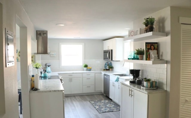 A modern kitchen is presented, featuring white cabinetry and a spacious countertop. Stainless steel appliances, including an oven and microwave, are visible. A window allows natural light to fill the room, and decorative elements, such as plants and wall art, enhance the inviting atmosphere.