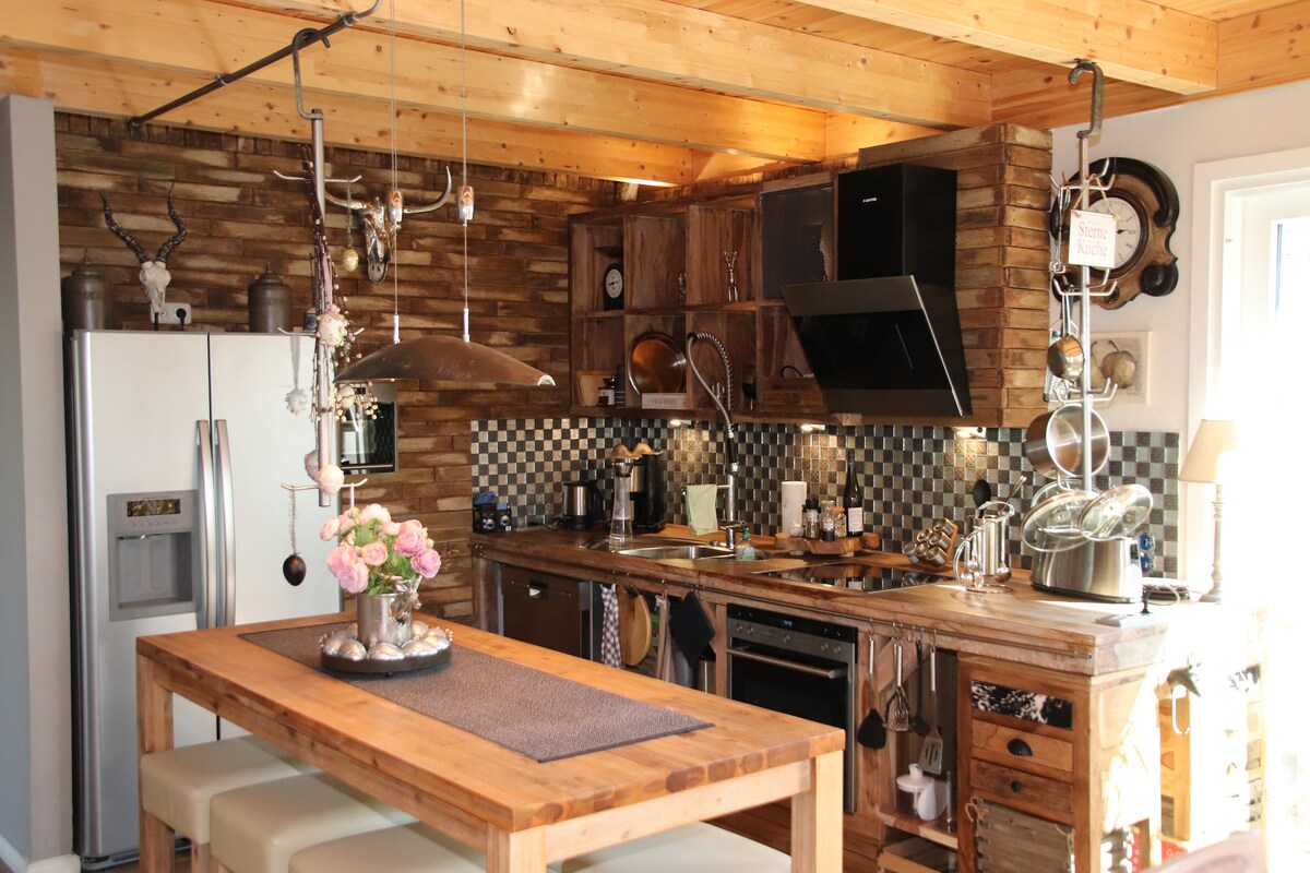 The kitchen showcases a blend of rustic wood finishes and modern appliances. A spacious wooden dining table is accompanied by upholstered seating. The arrangement includes a refrigerator, stove, and sink, all illuminated by natural light from windows, enhancing the functionality of the space.