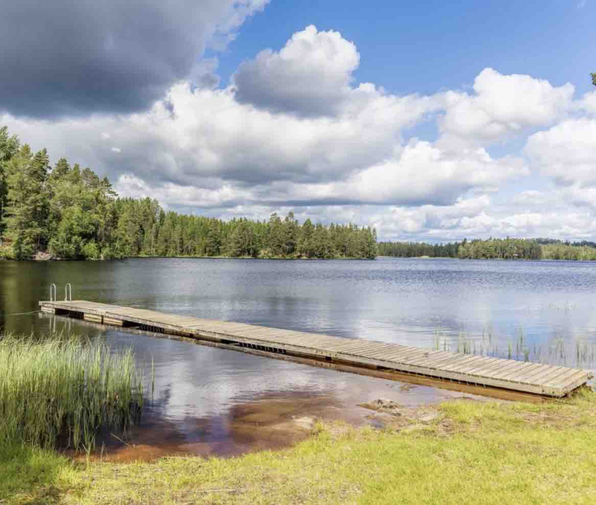 A wooden dock extends over calm waters, surrounded by lush greenery and trees. The lake reflects the sky, dotted with clouds, enhancing the tranquil atmosphere. The shoreline features patches of grass and sandy areas, providing a peaceful natural setting.