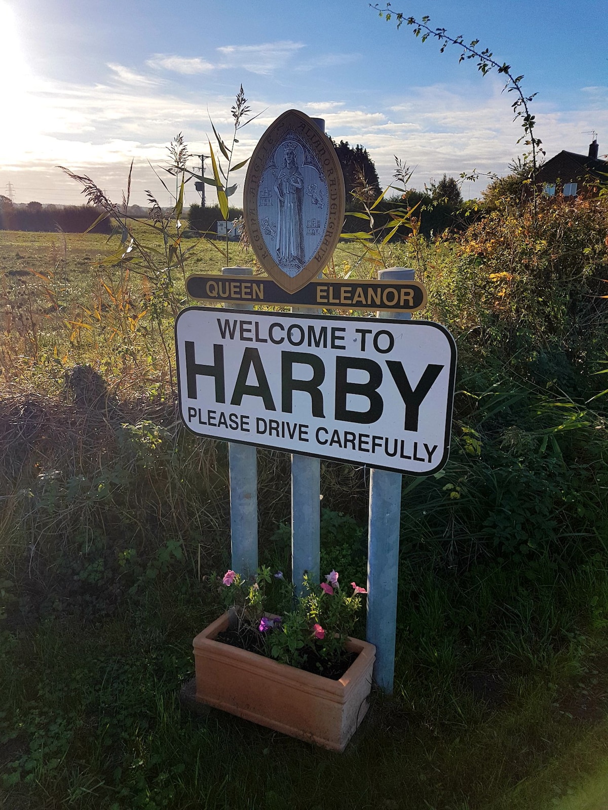 A welcome sign for the village of Harby is displayed prominently, featuring the name in bold letters. Below, a planter with blooming flowers adds a touch of color. The background reveals green fields under a clear sky with soft clouds.