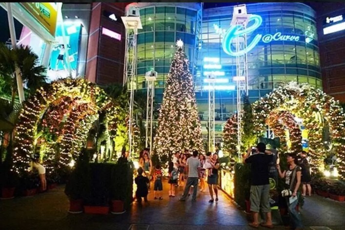 A vibrant outdoor shopping area is decorated for the holiday season. A large Christmas tree adorned with lights stands prominently, surrounded by festive foliage and illuminated archways. People of various ages can be seen enjoying the festive atmosphere, capturing photos amidst the twinkling lights.