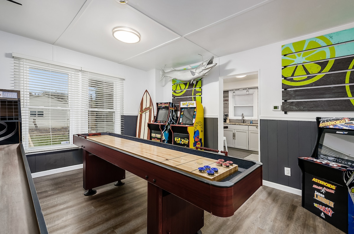 A game room is depicted, featuring a shuffleboard table in the center with colorful game chips nearby. Arcade machines line the walls, showcasing vibrant designs. Sunlight enters through a large window dressed with white blinds, illuminating the playful atmosphere created by the bright wall art.