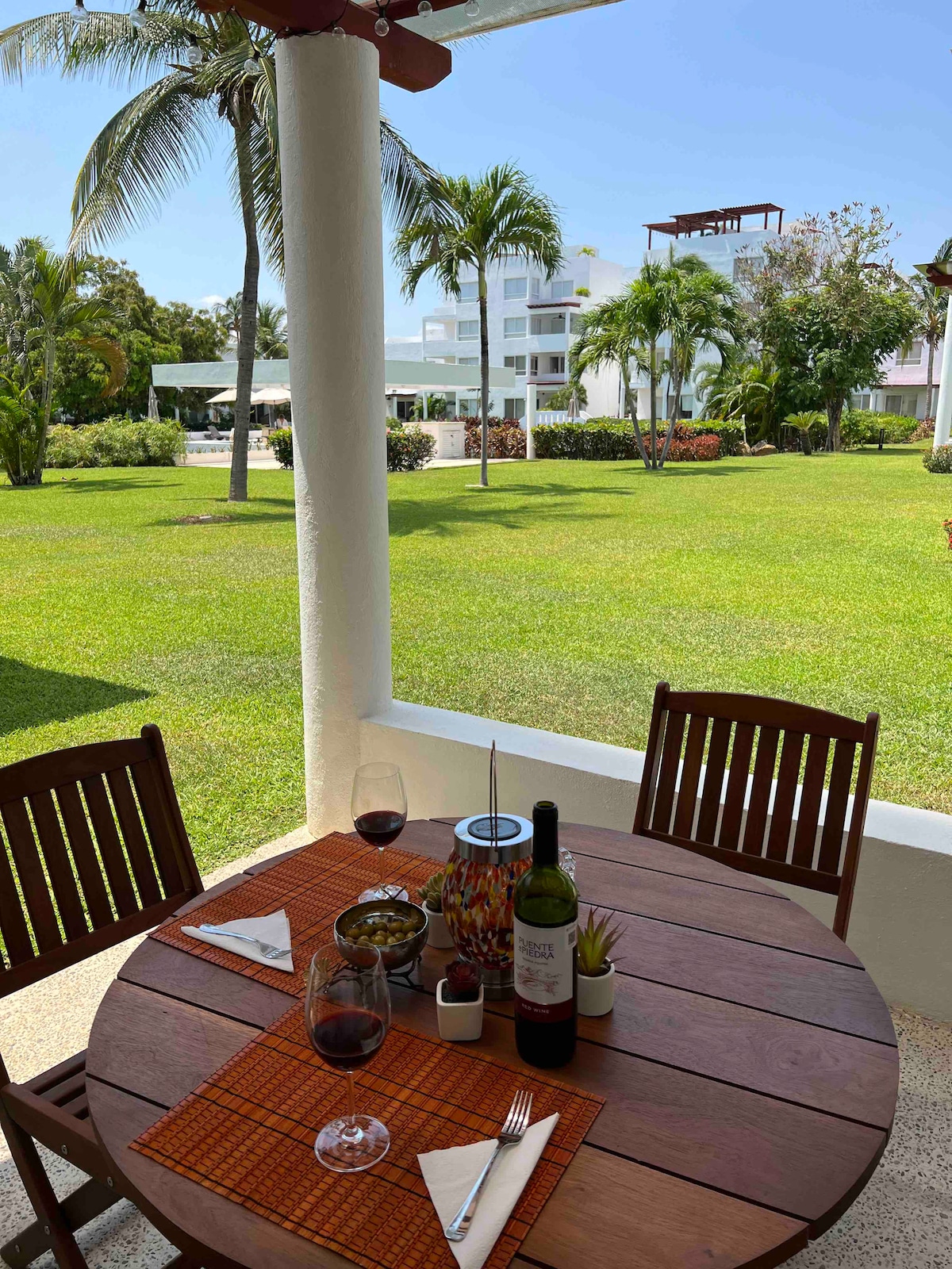A round wooden table is set for two, featuring wine glasses, a bottle of wine, snacks, and decor on a vibrant table runner. Lush green grass and tropical palm trees create a serene backdrop, with a view of the nearby residential buildings under a clear blue sky.