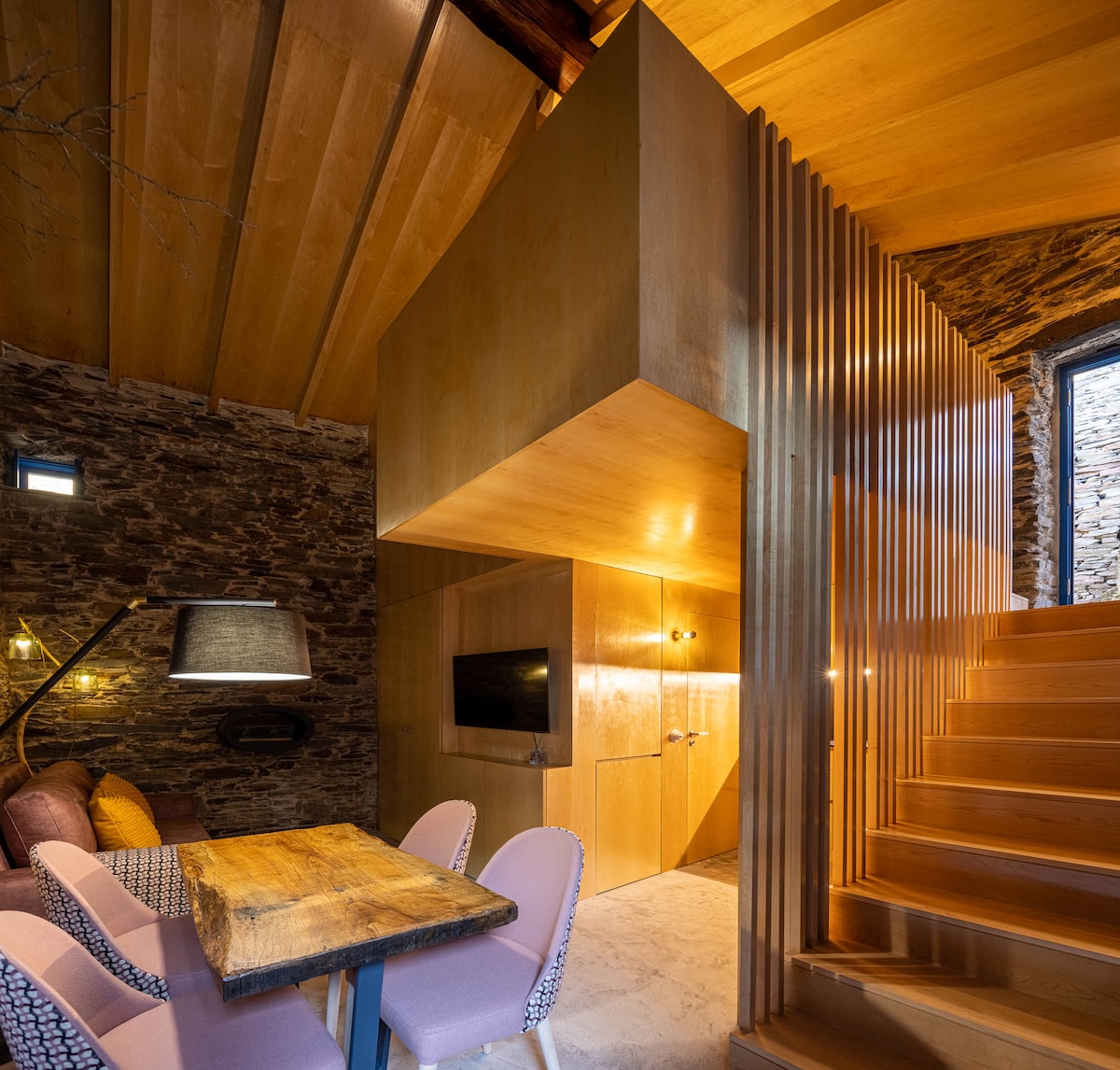 The image presents a warm interior featuring polished wood finishes and natural stone walls. A staircase leads to a mezzanine level, while a dining area with a rustic wooden table and upholstered chairs is visible. Soft lighting highlights the inviting ambiance.