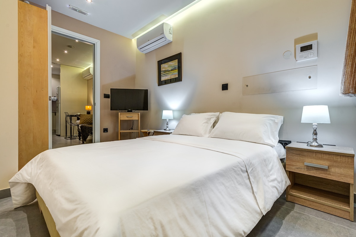 A spacious bedroom features a king size bed with crisp white linens. Nightstands with small lamps are positioned on either side of the bed. A wall-mounted television is visible across from the bed, and air-conditioning units are installed for comfort.