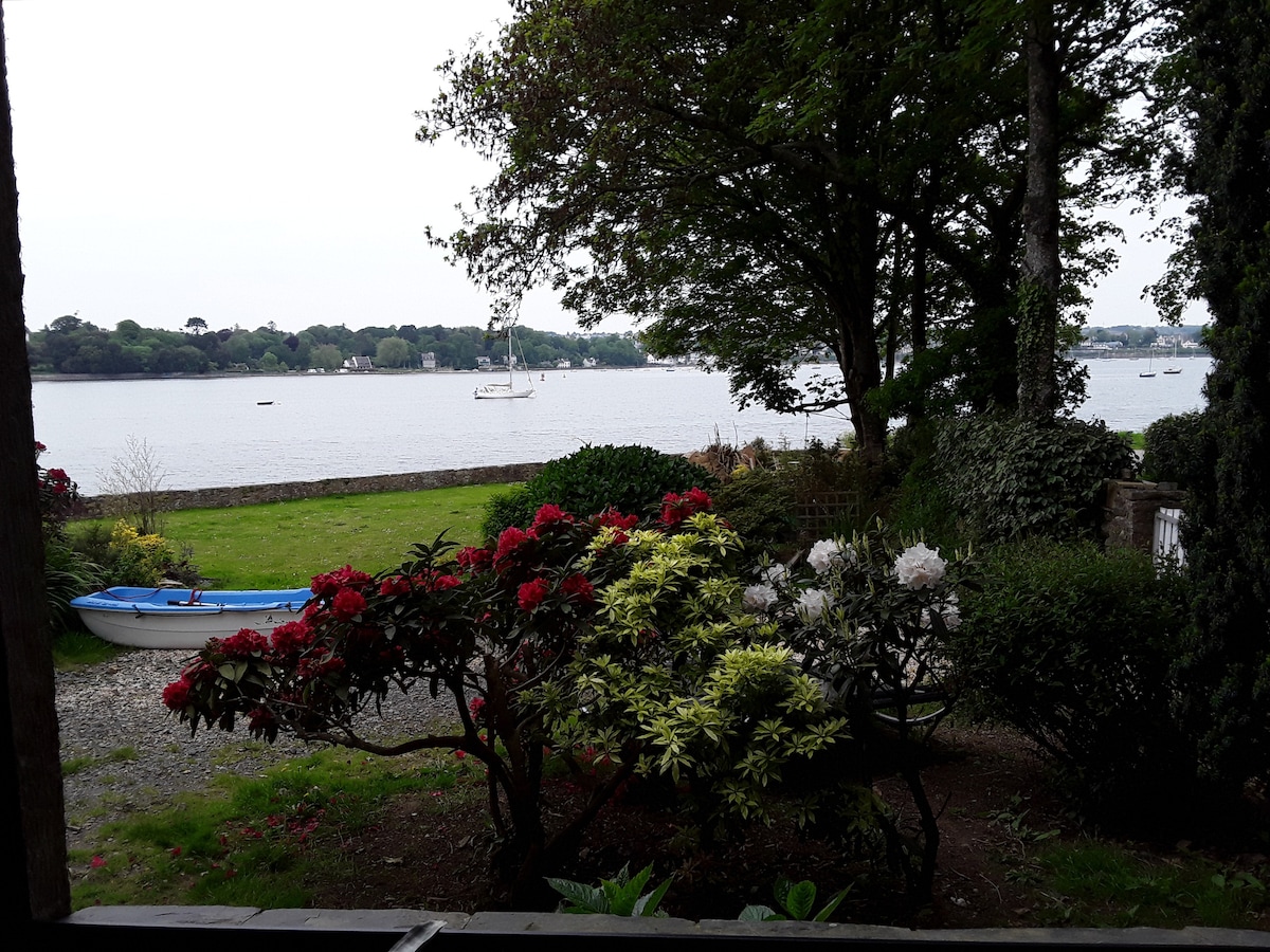 A serene view of the waterfront is showcased, with boats gently bobbing in the distance. Lush greenery and vibrant flowers in various colors frame the scene, creating a peaceful outdoor setting. A blue kayak is situated nearby, offering a glimpse of available water activities.