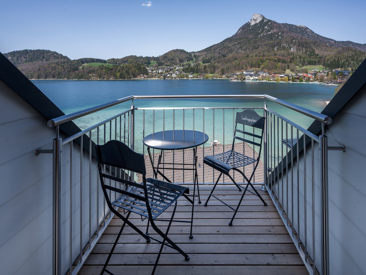 A small rooftop terrace features a round table and two black metal chairs, offering views of the tranquil lake and lush green hills. The serene backdrop includes a distant mountain under clear blue skies, creating a peaceful outdoor atmosphere.