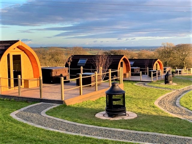 Several wooden cabins are arranged on a gentle slope, each featuring a private hot tub on the patio. Green grass surrounds the stone pathways leading to the cabins, with trees and open fields visible in the background under a bright sky.