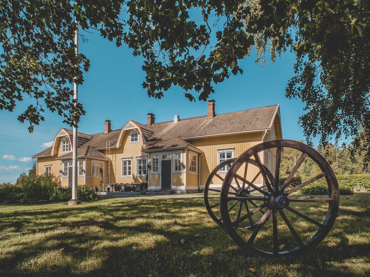 A well-preserved yellow wooden house features multiple gabled roofs and large windows, surrounded by greenery. A vintage wooden wheel is positioned prominently on the lawn, adding a rustic charm to the environment. Clear blue skies provide a bright backdrop to the inviting scene.