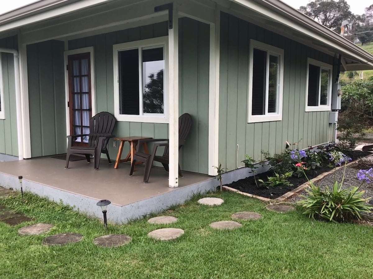 A charming exterior of the country home is shown, featuring a small patio area with two chairs and a side table. Manicured grass and garden beds frame the entrance, providing access to the home. Two large windows allow natural light into the interior.