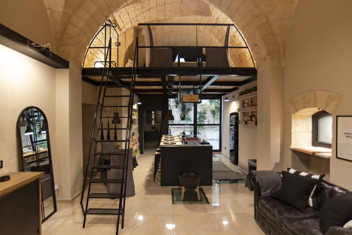 A modern loft space is characterized by high arched ceilings and open floor plan. A sleek kitchen island is positioned centrally, with a staircase leading to a lofted area. Large windows provide natural light, highlighting the stylish furnishings and minimalist decor.