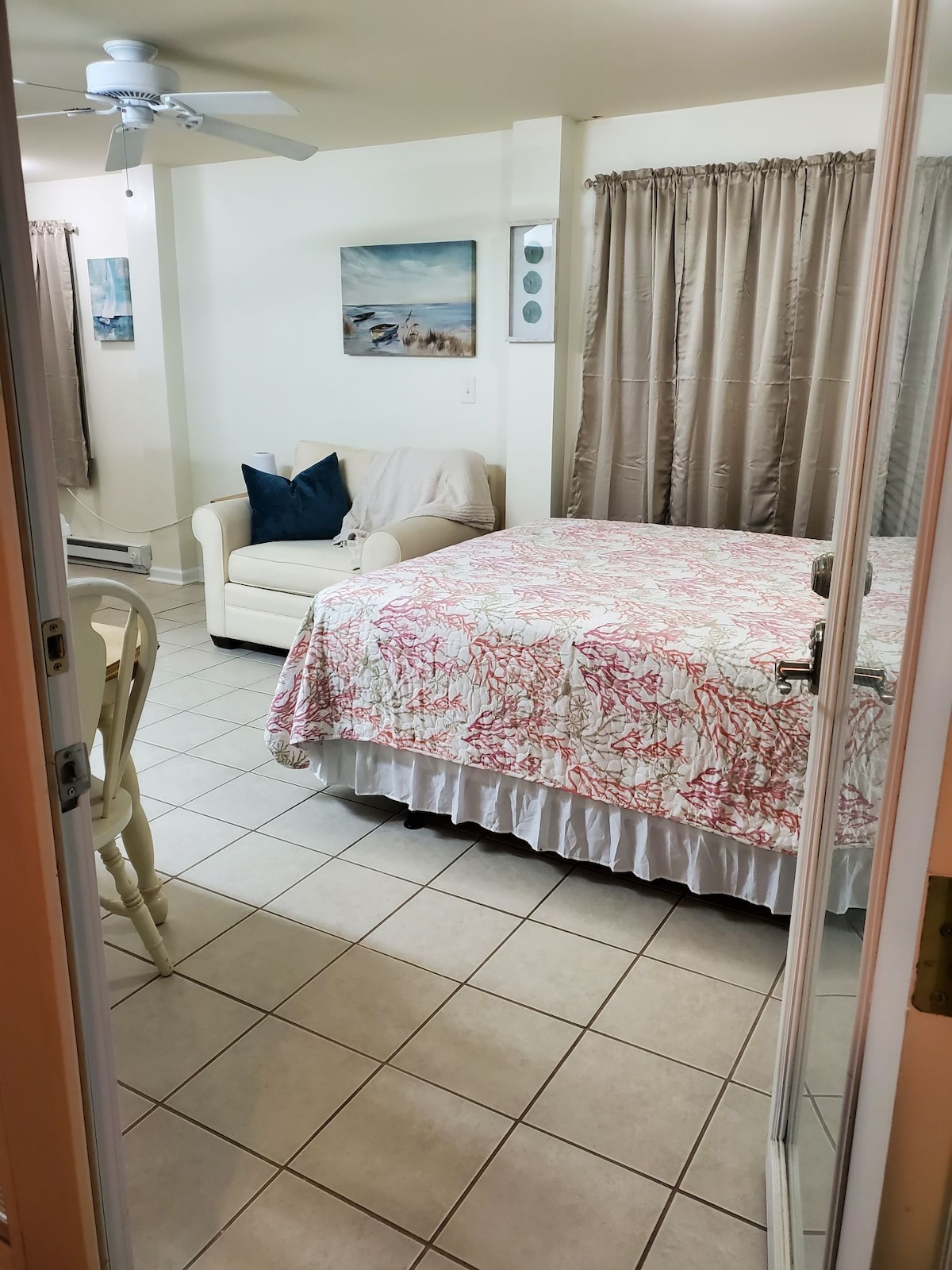 An inviting efficiency apartment features a spacious bed dressed in a floral comforter, complemented by a cozy seating area with a light-colored armchair. Soft curtains frame the windows, and wall art depicting serene beach scenes adds a touch of charm to the room.
