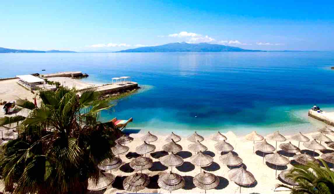 The beachfront scene features numerous thatched umbrellas arranged in neat rows on the sandy shore. Calm blue waters stretch into the distance, reflecting a clear sky dotted with a few clouds. Faint mountainous landscapes are visible on the horizon, creating a natural backdrop.