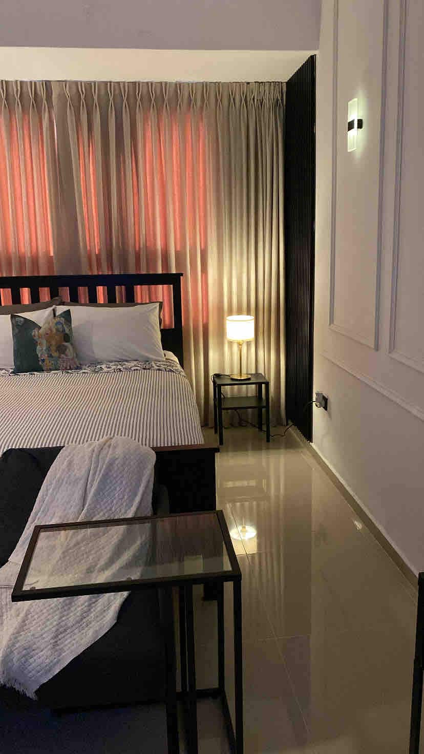 A cozy sleeping area features a dark wooden bed dressed with patterned linens and a decorative pillow. Soft, warm light filters through sheer curtains, illuminating a bedside table with a lamp. A throw blanket is casually draped over a low table, enhancing the inviting ambiance.