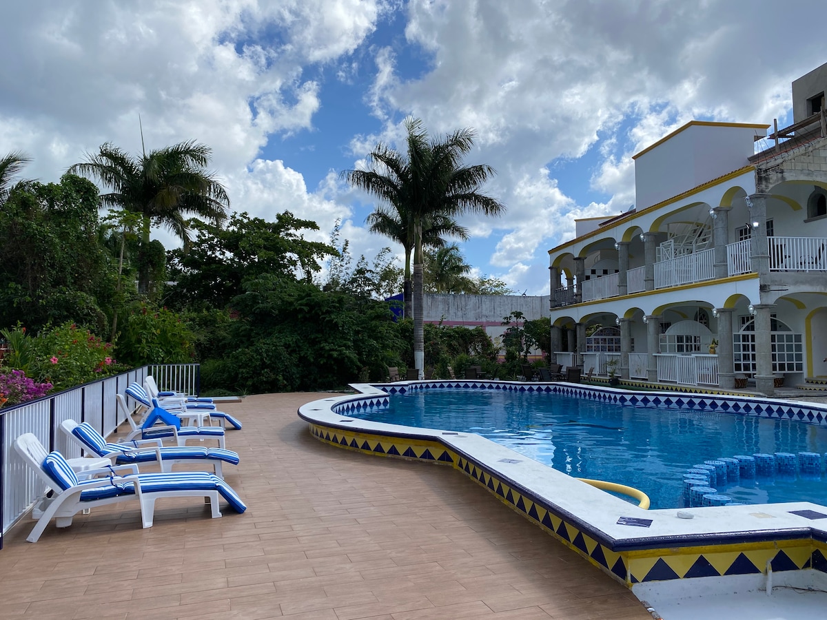 A private outdoor area features a large swimming pool surrounded by inviting lounge chairs. Lush greenery and blooming flowers are visible in the background, along with a multi-level villa that showcases arched balconies and white railings. The sky is partly cloudy, creating a serene atmosphere.