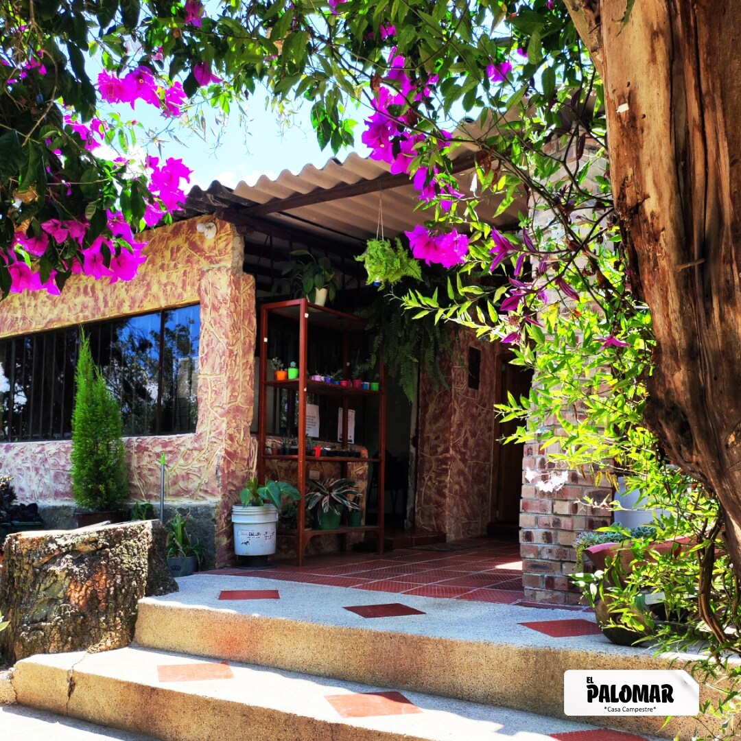 The entrance features a charming facade adorned with vibrant purple flowers. A stone pathway leads to the door, where an open patio extends with greenery and decorative plants visible. The structure showcases a mix of natural elements, inviting guests into a tranquil setting.