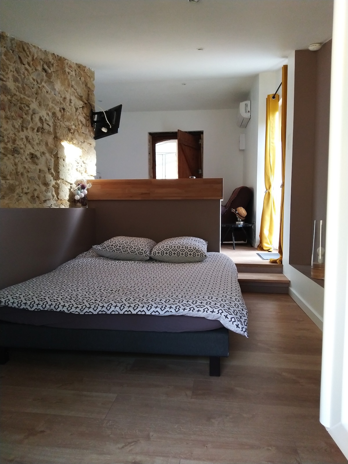 A cozy sleeping area is presented with a spacious bed featuring patterned linens. Natural light filters through a nearby window, illuminating the warm tones of the stone wall and neutral-colored walls. A doorway leads to another room, enhancing the sense of openness.