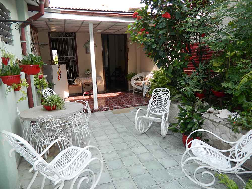 A tranquil outdoor area features a tiled patio surrounded by lush greenery and vibrant flowers. Four white wrought-iron chairs are arranged around a circular table, inviting relaxation. An entrance to the interior is visible, framed by a canopy providing shaded comfort.