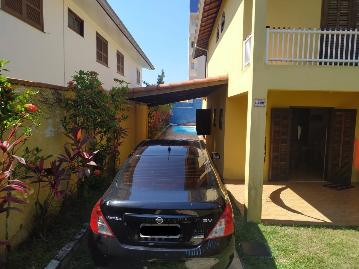 A car is parked under a canopy, providing protection from weather elements. The driveway leads towards a pool area visible in the background, surrounded by greenery and flowers. The exterior walls of a two-story building are painted in a light yellow tone, contrasting with the vibrant vegetation.