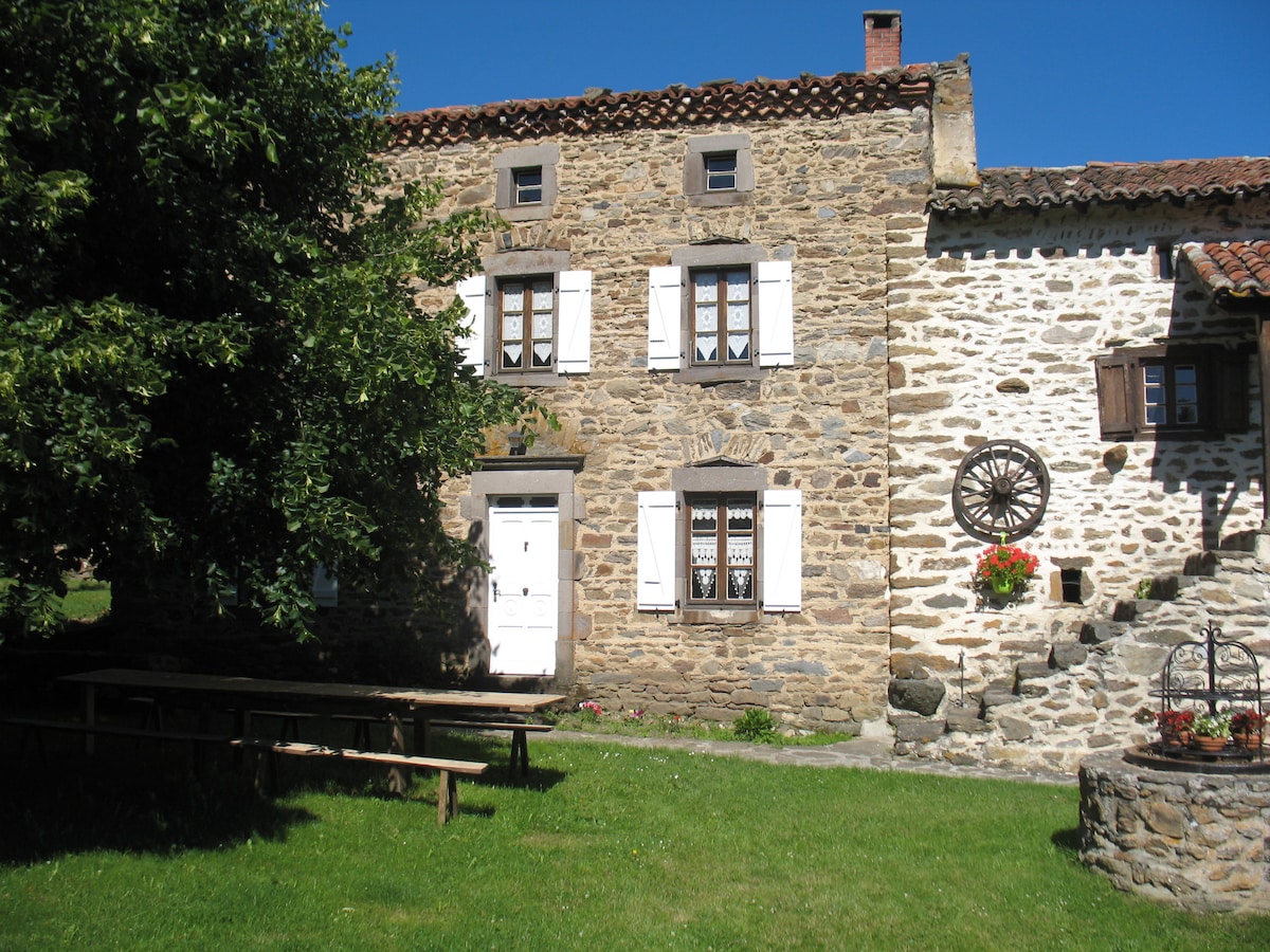 A stone structure characterized by a rustic facade features white shutters and a door accented with decorative lattice. The grassy area in front accommodates a long wooden bench, while a pathway lined with flowers leads to a charming stone building, enhancing the natural setting.