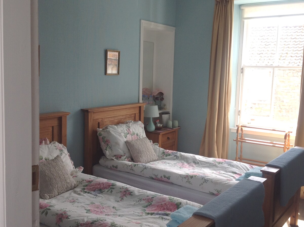 A tranquil twin bedroom features two comfortable single beds, adorned with floral bedding. Light blue walls complement the serene atmosphere. Natural light enters through a large window, which is dressed with soft curtains. A mirror and wooden furnishings add to the room's welcoming ambiance.