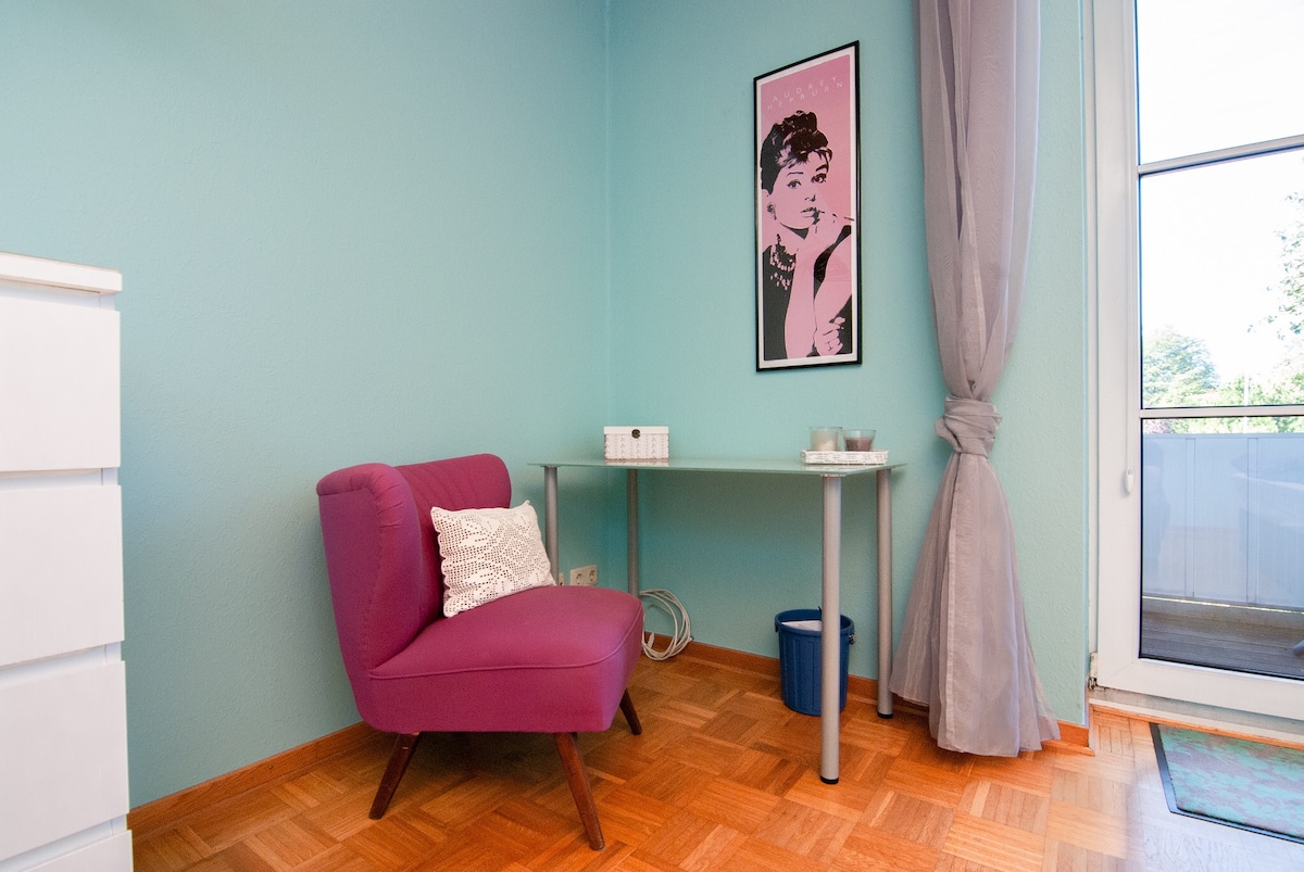 A bright corner of the room features a pink armchair with a textured pillow beside a simple table. A small wastebasket is visible below the table, and natural light streams through the glass doors leading to a balcony, adorned with light curtains.