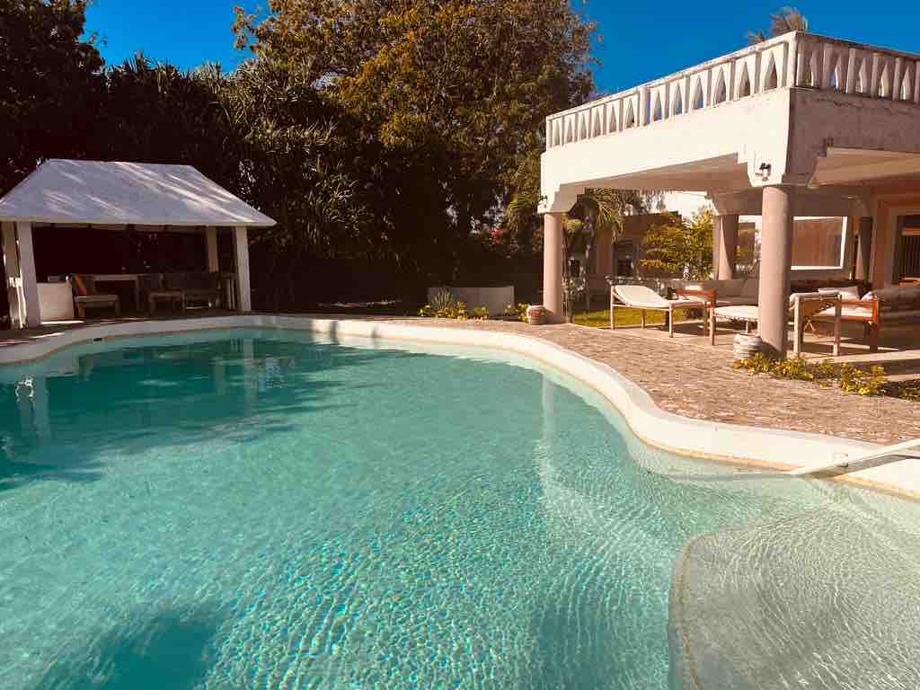 A tranquil pool area features clear blue water reflecting sunlight. Surrounding greenery adds to the natural ambiance. A shaded seating area is positioned beside the pool, providing a space for relaxation under the open sky.