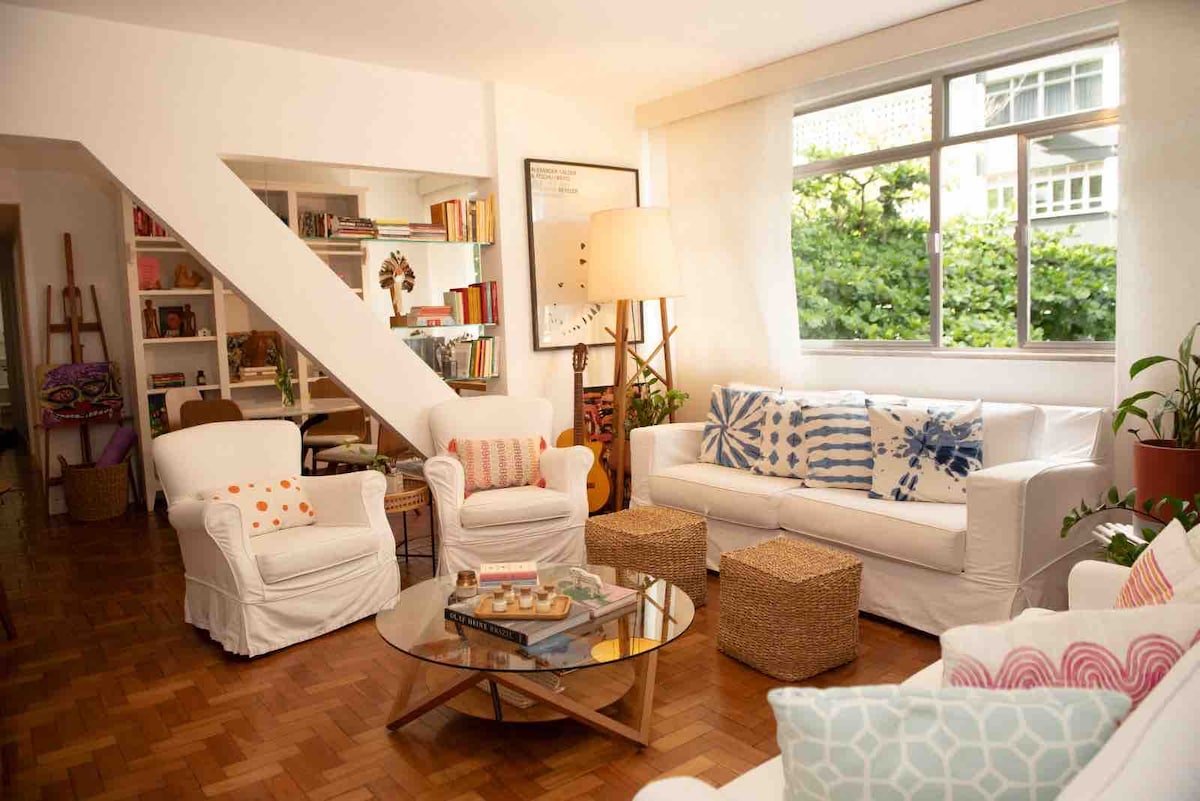 The living area features comfortable white armchairs and a sofa, adorned with patterned cushions. A circular glass table sits at the center, surrounded by natural light filtering in from a large window. Bookshelves are filled with books, and greenery can be seen outside.
