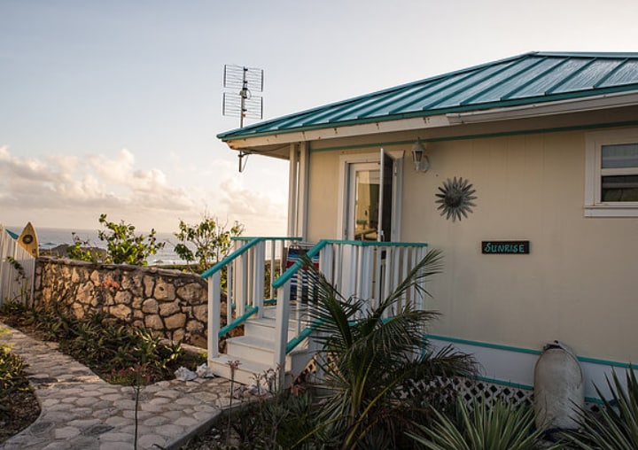 A charming exterior view of the studio cottage is presented, featuring a turquoise-accented ramp leading to the entrance. The light-colored siding is complemented by a metallic roof. Surrounding greenery and a stone pathway enhance the inviting ambiance of the property.