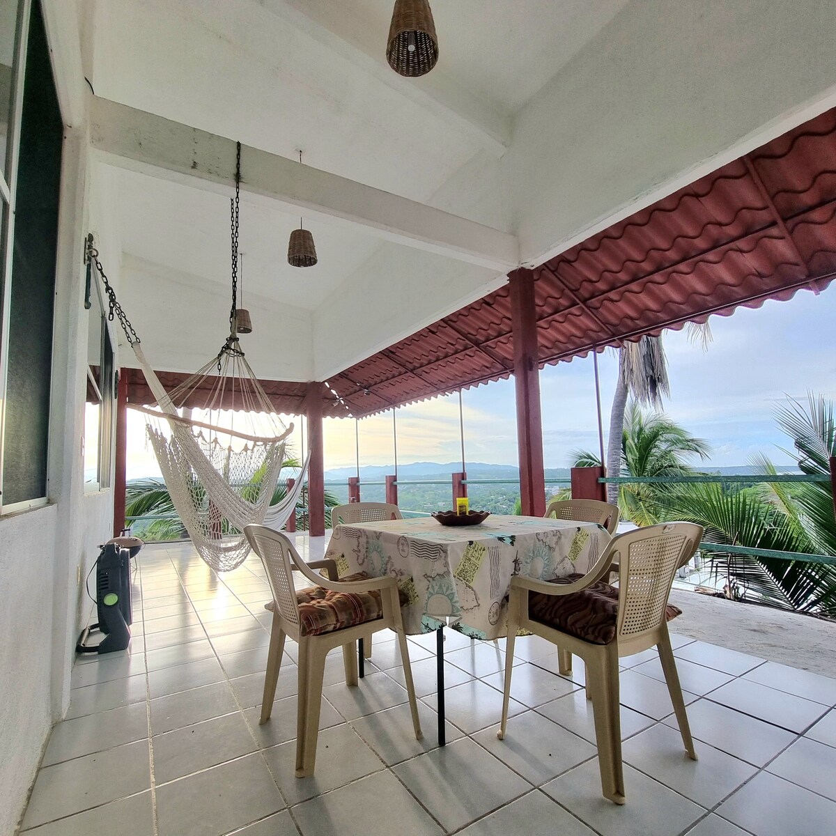 A spacious terrace is shown with a dining table surrounded by chairs, ideal for enjoying meals outdoors. A hammock is suspended nearby, complemented by palm trees in the background. The area is well-lit with natural light, offering sweeping views of the surrounding landscape.
