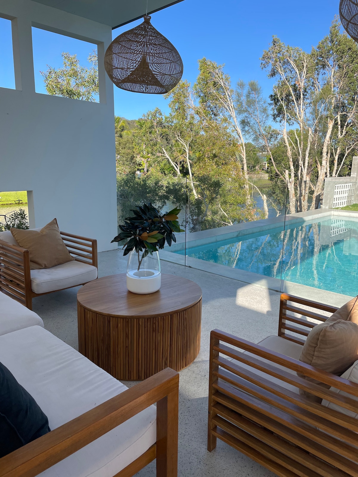 A comfortable seating area is presented with a round wooden table and two wooden armchairs, adorned with cushions. Large glass windows open to views of lush trees and a tranquil pool, reflecting the clear blue sky, creating a serene indoor-outdoor atmosphere.