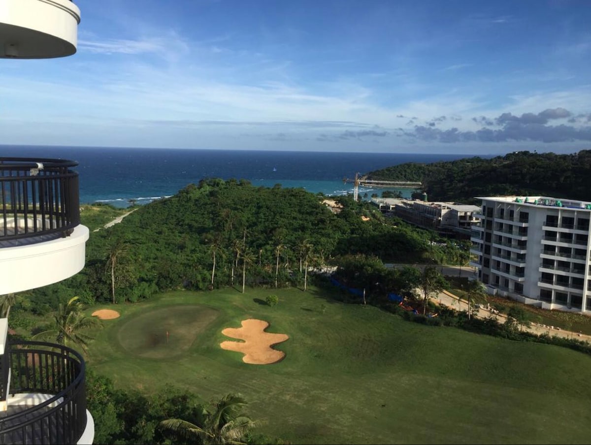 An expansive view of lush green scenery transitions into the tranquil blue ocean. The golf course, with its sandy patches, is visible below the balcony. The landscape is framed by palm trees, while the coastline is dotted with gentle waves under a clear sky.
