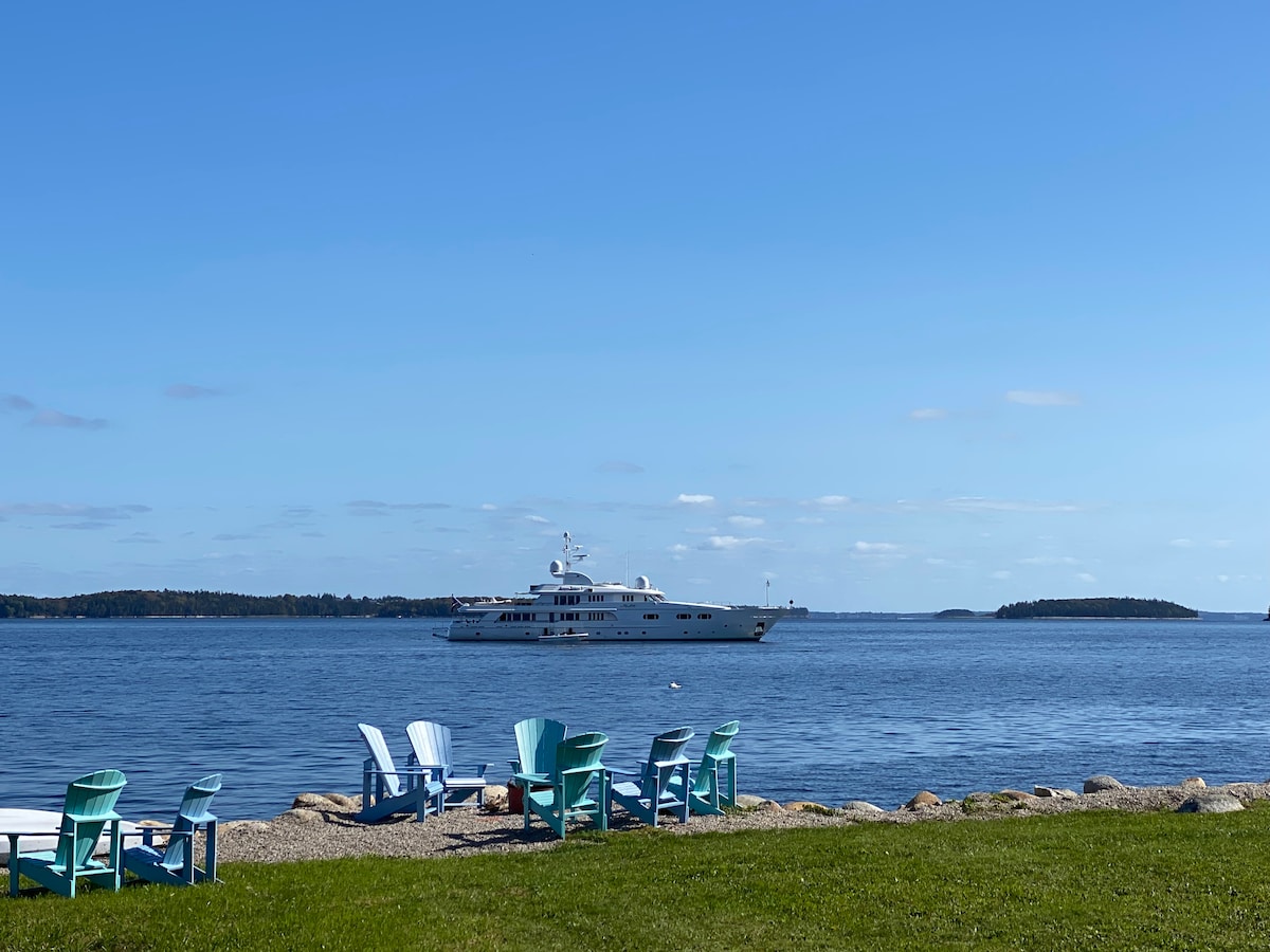 A calm ocean scene features a large yacht gliding across the water, with lush islands visible in the distance. In the foreground, several light blue Adirondack chairs are arranged on the grassy shoreline, inviting relaxation and scenic views of the coastline.