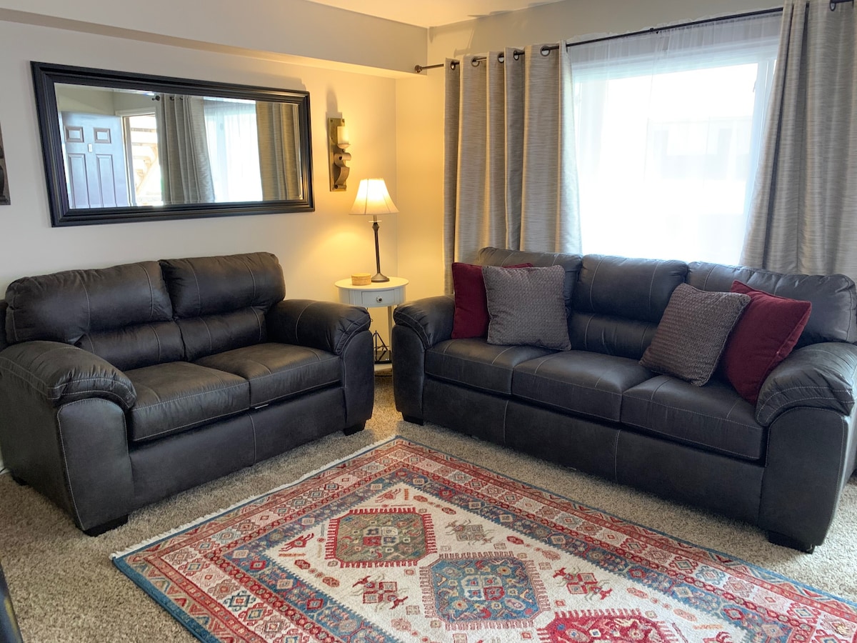 A comfortable living area is furnished with two dark leather sofas, positioned across from each other. A patterned area rug lies beneath the seating, and a lamp stands beside a small table. Natural light filters through the large windows adorned with soft curtains.