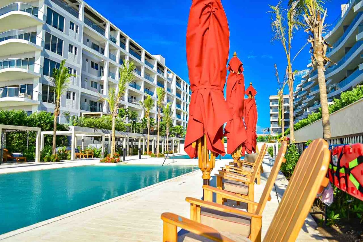 Lounge chairs with bright umbrellas are positioned alongside a shimmering swimming pool. The background showcases a modern building with multiple stories and palm trees lining the pool area, creating a relaxing environment for guests.