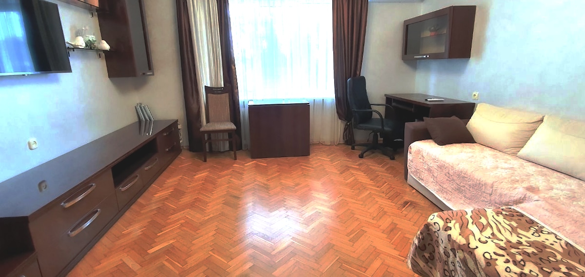 A living area features wooden flooring and large windows allowing natural light. A cozy sofa, a desk with a chair, and a small table are arranged neatly. Dark curtains frame the windows, and modern storage units provide a functional space.